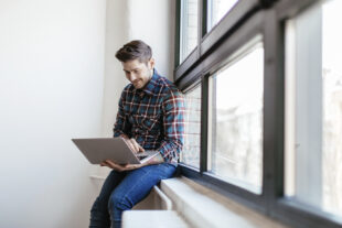 Man sat on a window ledge, using a laptop and smiling