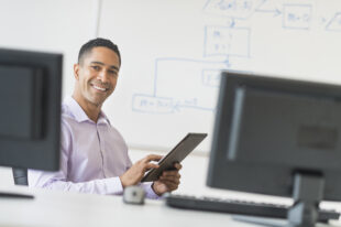 Smiling businessman holding a digital tablet, sitting at a desk with computers on