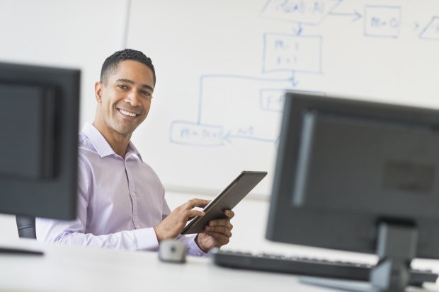 Smiling businessman holding a digital tablet in front of a desk with computers on