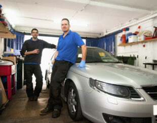 Two mechanics in a garage, leaning against a car