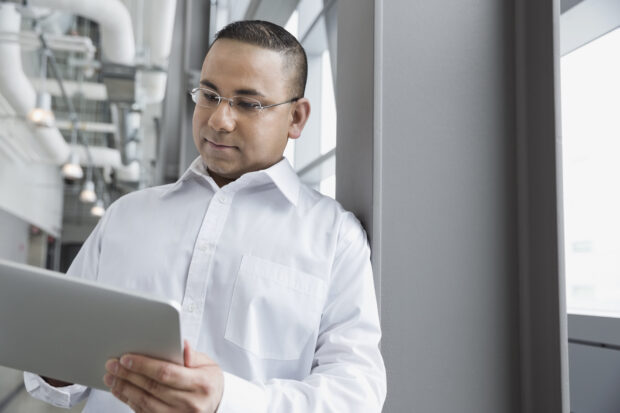 Man leaning against a workplace wall looking at a tablet device