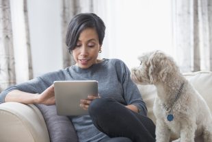 Woman sitting on a sofa, looking at a digital tablet, with a dog looking on